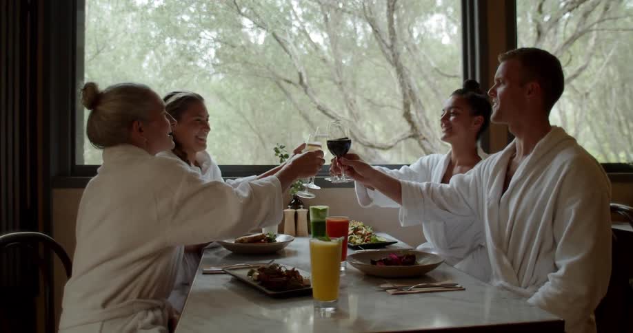 Four people in white bathrobes sit around a dining table and all raise their glasses to toast