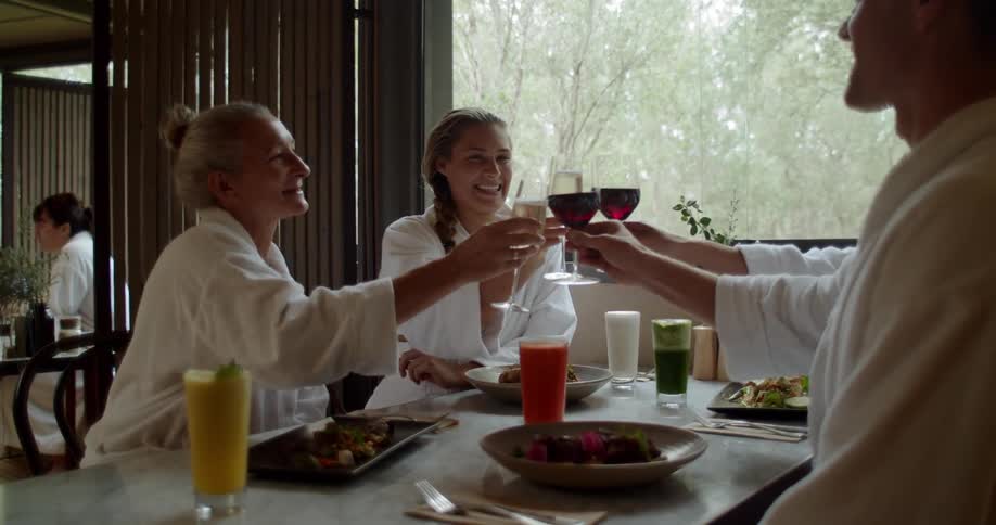 A group of people in white robes and seated at a dining table all hold up their glasses and toast each other