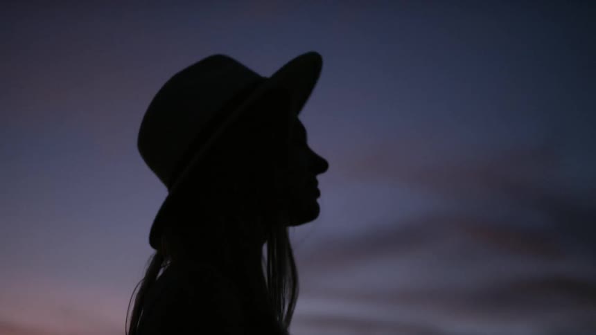 A woman wearing a hat turns her head as she looks at the sky at sunset