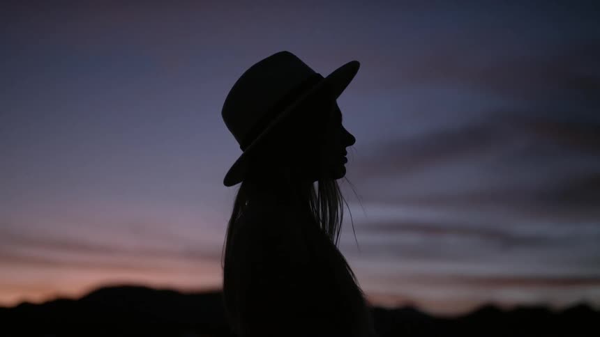 The silhouette of a woman in a hat standing outside at sunset