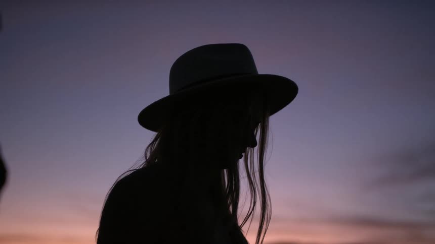 A woman in a hat is silhouetted against the sky at dusk