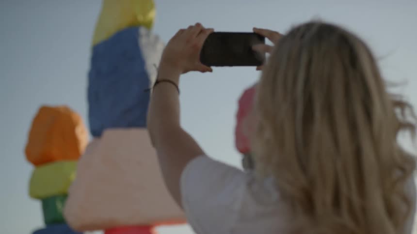 A woman holds up a cell phone to take a picture of rocks painted with bright colors