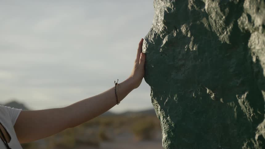 A white woman wearing sunglasses reaches out and touches a large rock in the desert