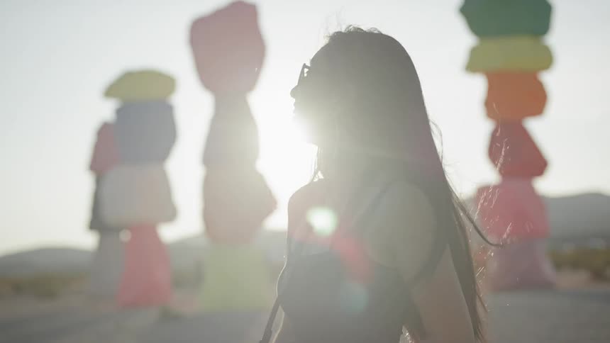 A young asian woman smiles as she stands outside in the sunlight near some brightly colored stacks of boulders