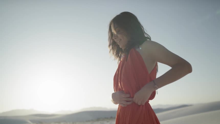 A brunette woman in a dress laughs while walking in the desert
