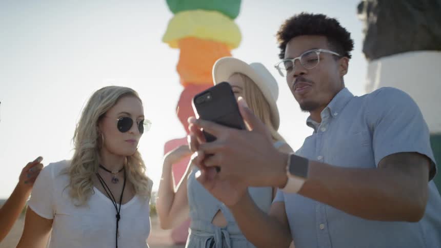 Three women and a man take a picture on his cell phone with large colorful sculptures in the background