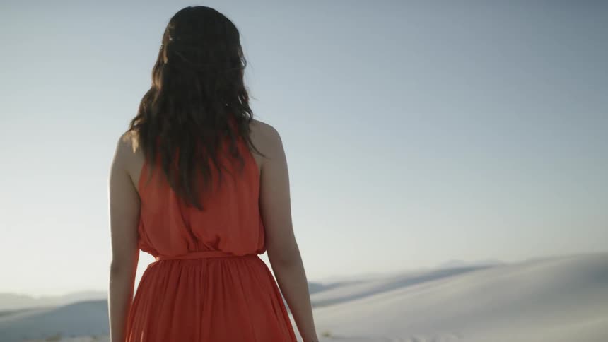 Brunette woman wearing a red dress stands on the white sand and looks around at sunset
