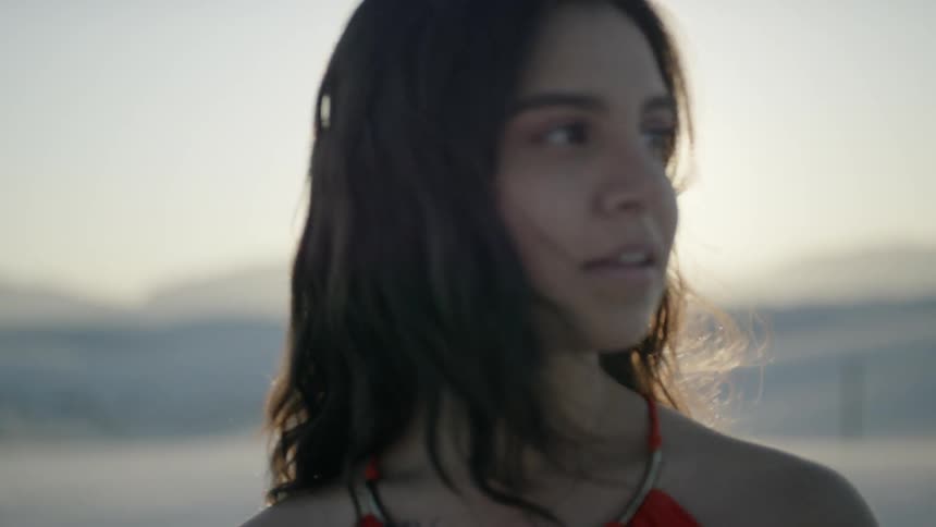 A woman looks around at the desert sand with the sun shining around and behind her