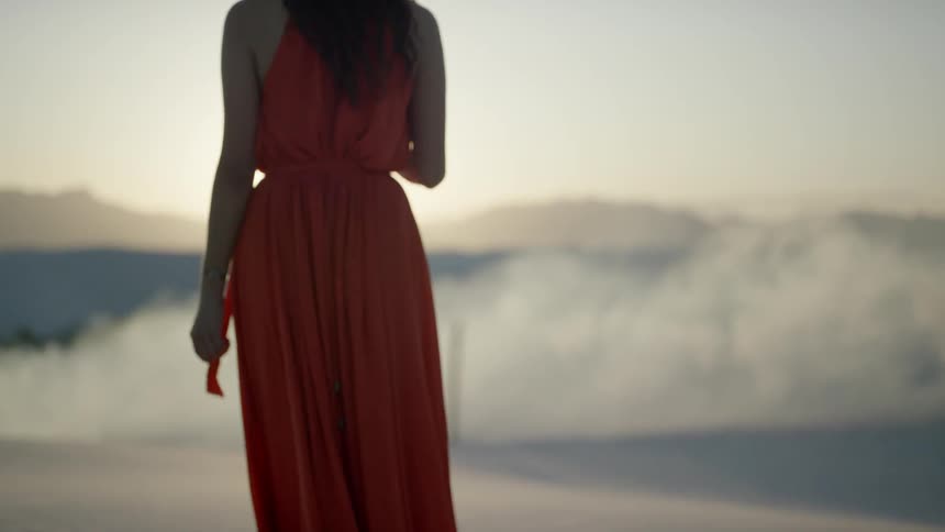 A young woman in a long orange dress walks outside while a large column of smoke is rising up in front of her