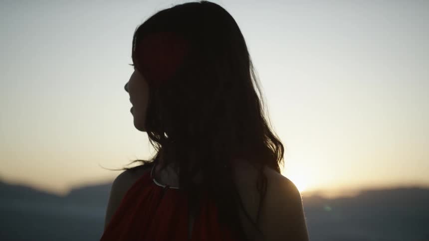 A woman in a red dress turns and looks at the desert sand as the sun sets
