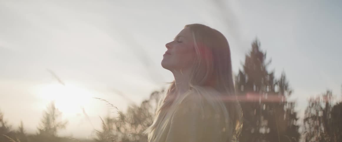 A blonde haired woman stands with her head up and eyes closed while taking in the sunset