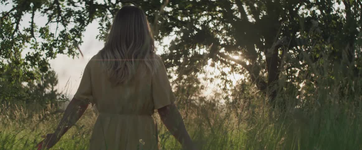 A woman in a full length dress walking through a field touching the tall grass at low sun