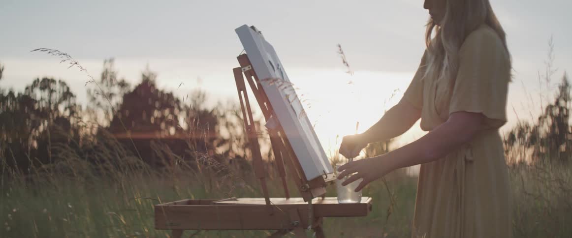 Woman paints standing among tall grasses in field at sunset