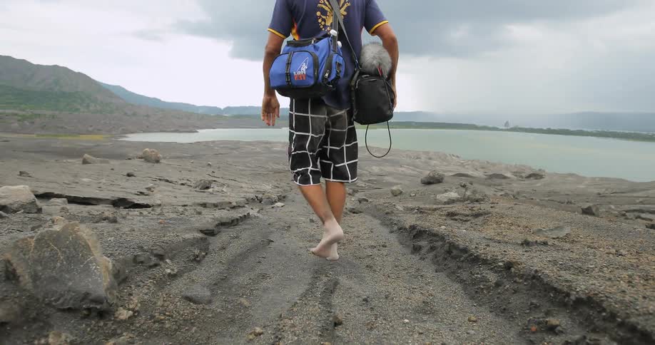 A man carrying several bags walking along a rocky beach toward water on a cloudy day