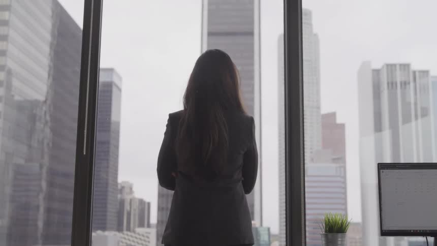 A woman in business attire stands and looks out an office window
