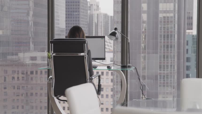 A woman is sitting at a modern desk with a glass top near some windows in a high rise building