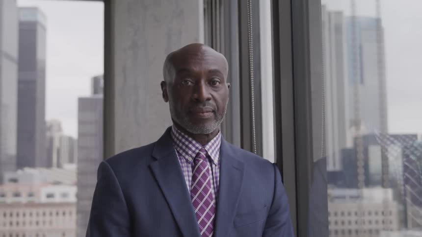 A man in a business suit standing in a corner office with windows turns his head