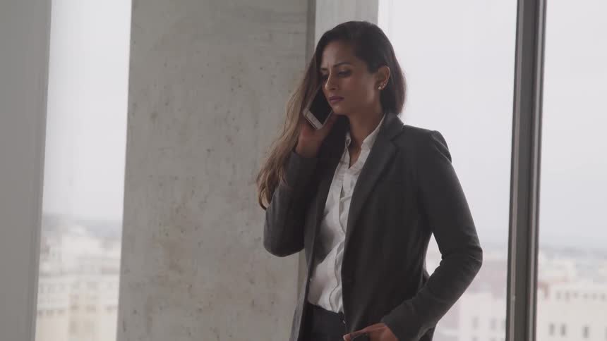 A woman in a suit standing near the windows in a skyscraper while holding a smartphone to her ear