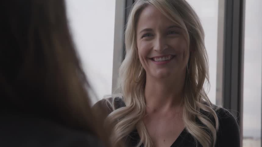 Blonde woman and brown hair woman look and smile at each other in a room