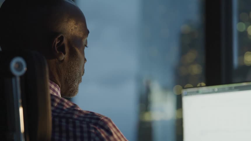 Bald headed black man sitting at the desk uses computer in the office at dusk