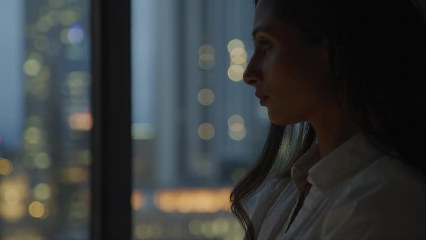 A woman stares out of large windows to the dark lights of the city