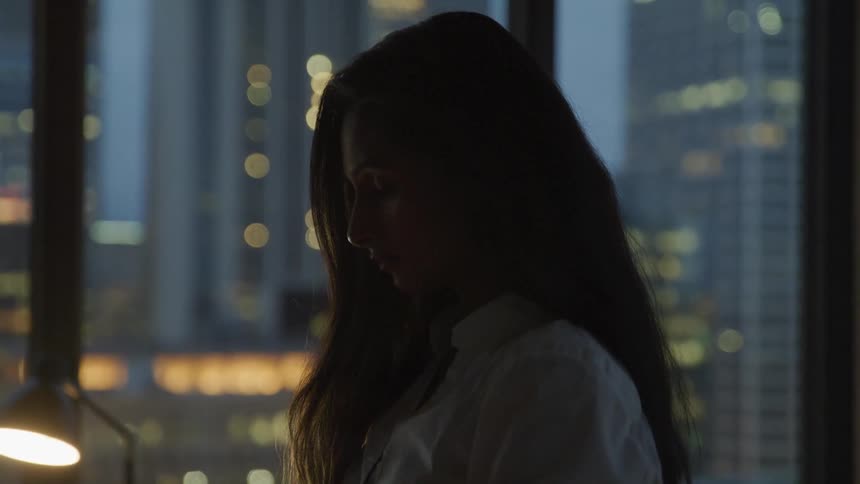 Businesswomans silhouette stands in the office with the view of tall buildings at dusk