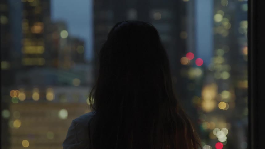 A woman standing staring out a window with buildings in the background