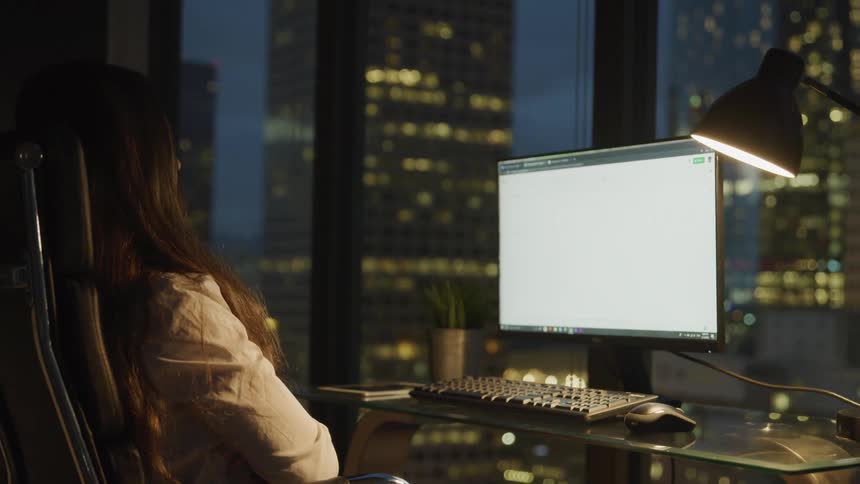 A young woman sits at her desk and looks at her computer screen