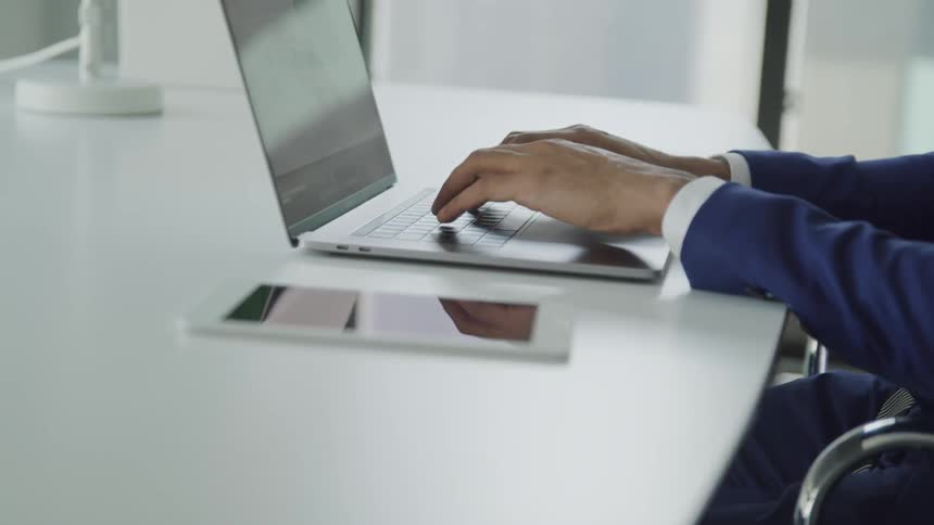 Male types on a laptop computer that sits on a white desk in an office