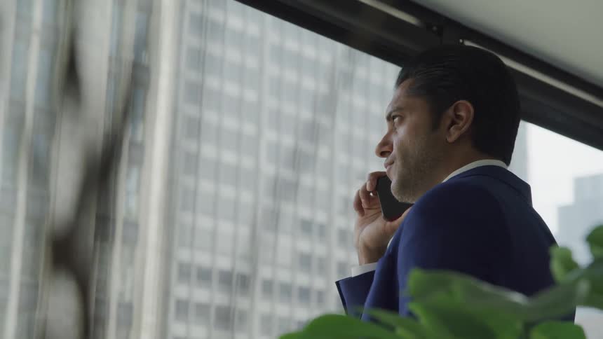 Businessman wearing a blue suit talks to someone on the phone looking out the window in the office