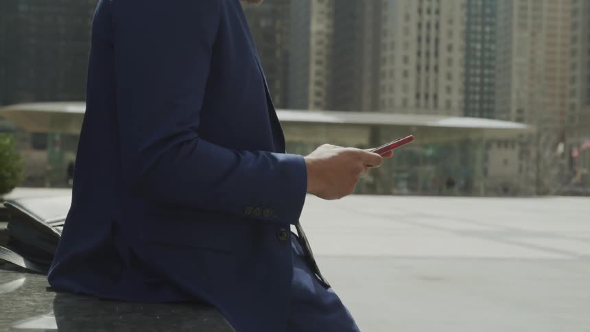 A man sits on a ledge outside and looking at his cell phone