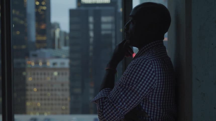 A man leans against the wall in an office and looks out the window with his hand on his chin