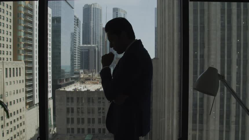 A man thinks while standing next to his office window in a city center