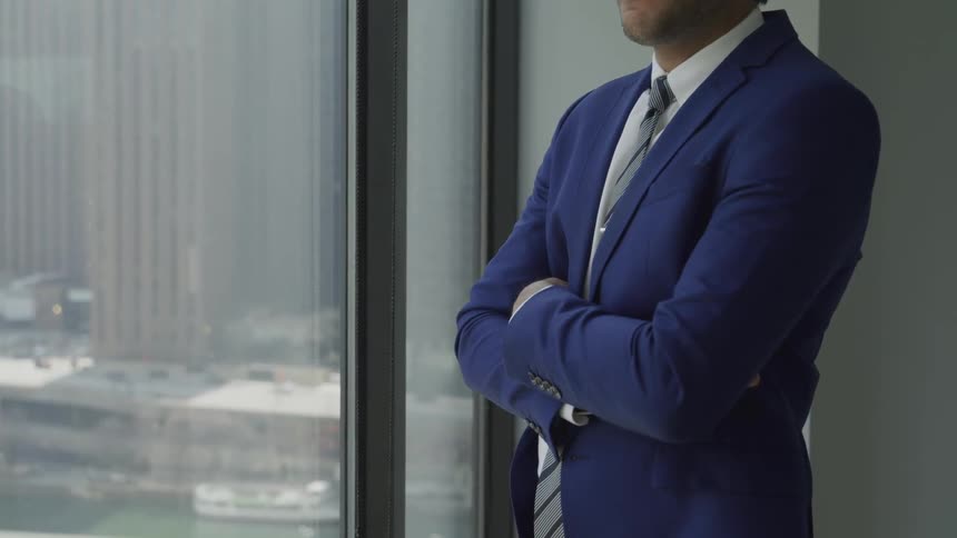 Man in suit stands with arms crossed next to window