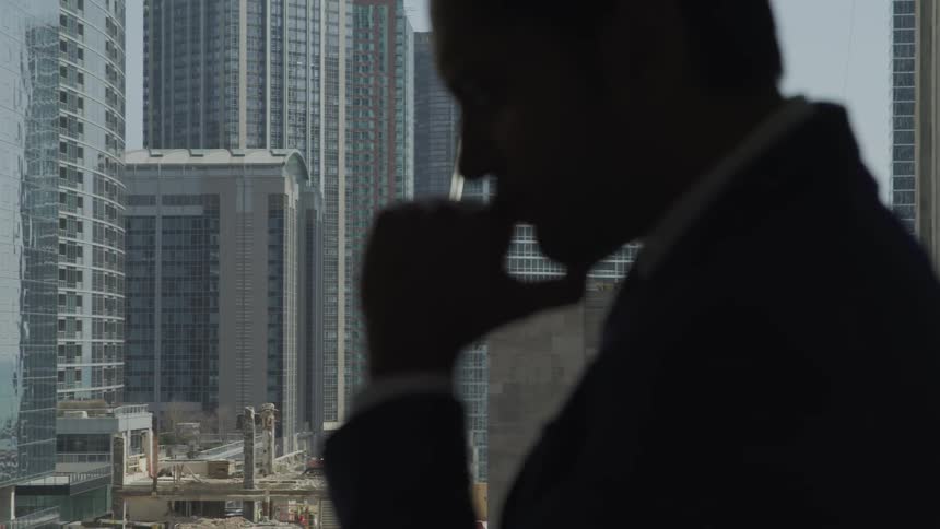 A man stands in front of a large window and rests his chin in his hands