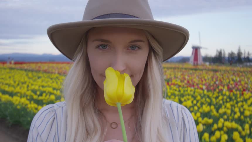 A woman wearing a hat is smelling a flower while standing in a field