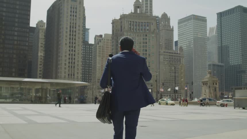 A man in business attire with a leather laptop case over his should holds his phone to his ear as he walks through an open plaza in the middle of a metropolitan area