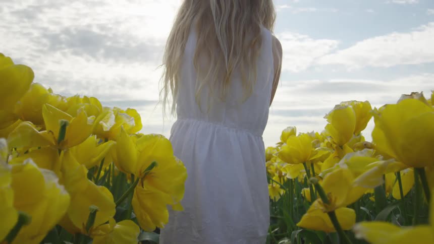 A white female in a white dress walks through a field full of yellow flowers