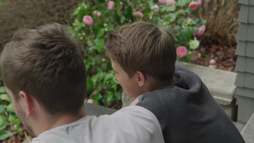 A young boy is hanging out with an older male outside near a garden as he chats and smiles
