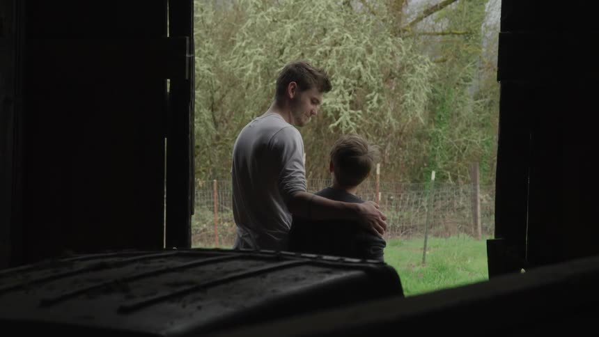 Two male siblings stand at the doorway to an open shed while looking out at the yard nearby