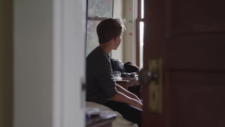 Boy sitting on the bed looks out the window in his room