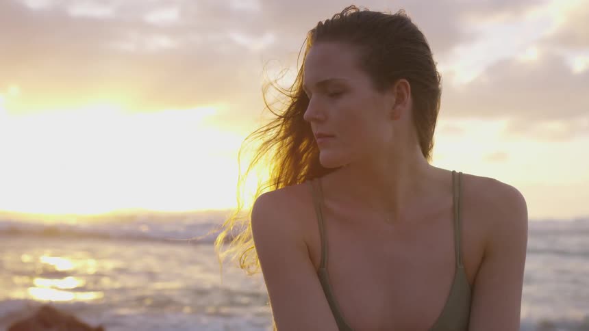 Brown haired woman in swimsuit looks sideways on the beach next to the sea at sunset