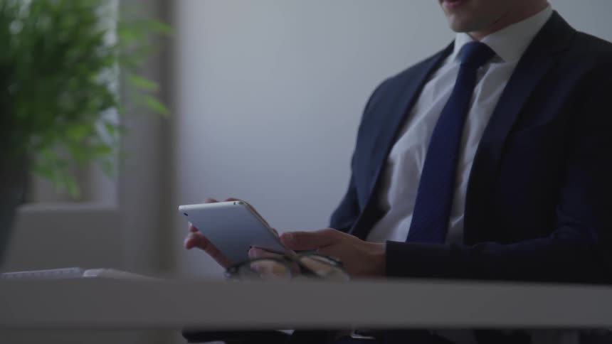 A man in a suit sits at a table tapping on a tablet