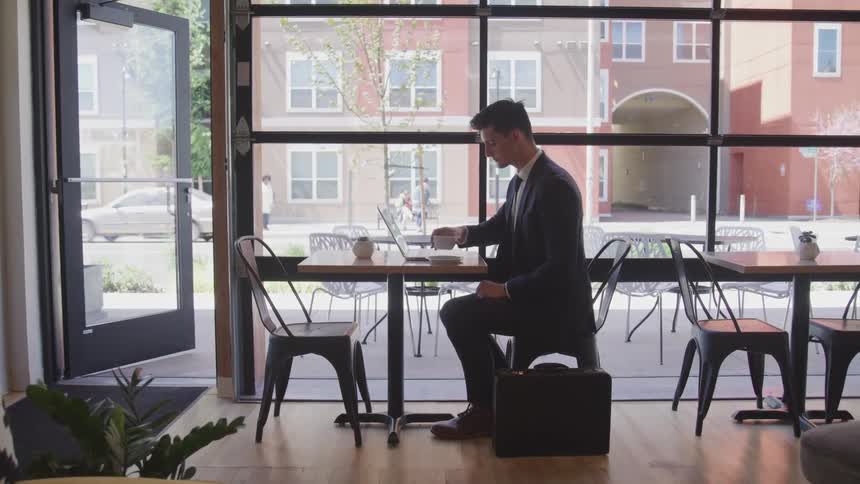 Businessman sitting at the table drinks coffee and uses laptop in the cafe