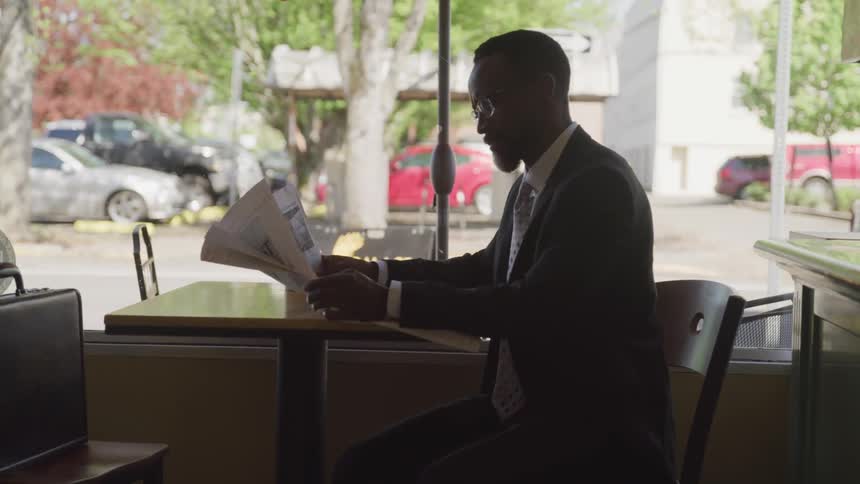 A black man sits at a table and reads a newspaper