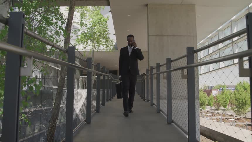 A black man in a suit talks on the phone while walking on an elevated walkway