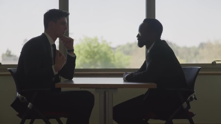 Two men sit at a small table next to a window while having a conversation