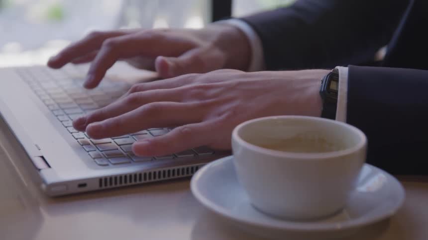 Male types on a laptop computer and drinks a cup of coffee while sitting at a table next to the window of a restaurant