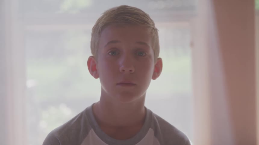 A boy standing with his back to a window in a room stares straight ahead