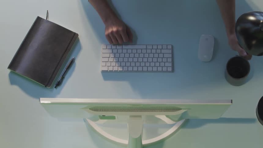 Birds eye view of a woman at a desk typing on her keyboard and drinking coffee
